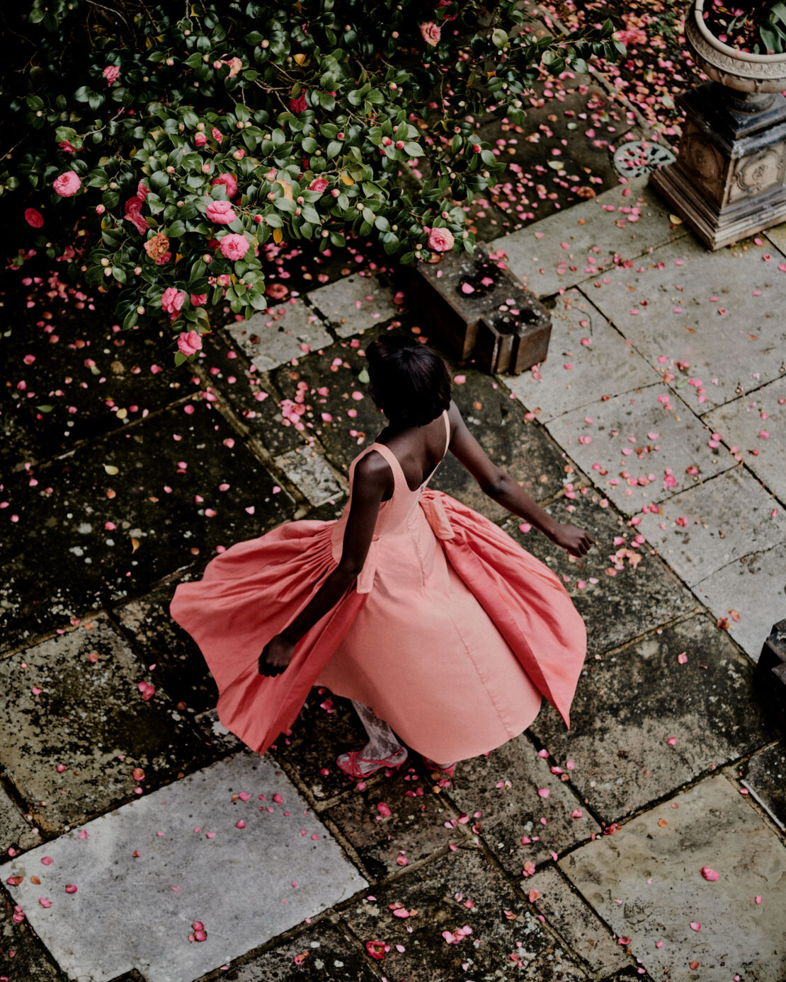 Overhead view of a Black woman in a pink dress surrounded by pink flower petals on a stone courtyard near green foliage and a stone urn.