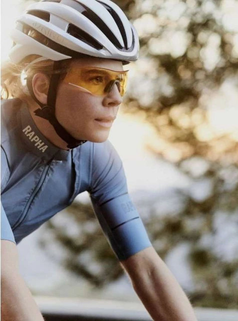 Close-up of a cyclist wearing a helmet and sunglasses, sporting a light blue cycling jersey with a blurred nature background.