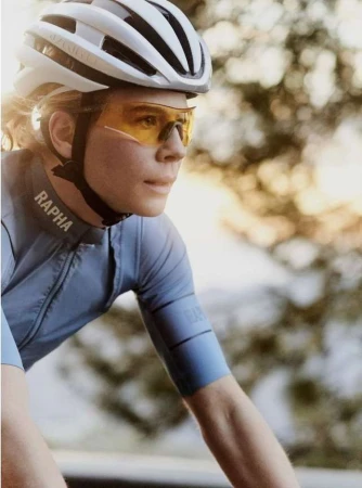 Close-up of a cyclist wearing a helmet and sunglasses, sporting a light blue cycling jersey with a blurred nature background.