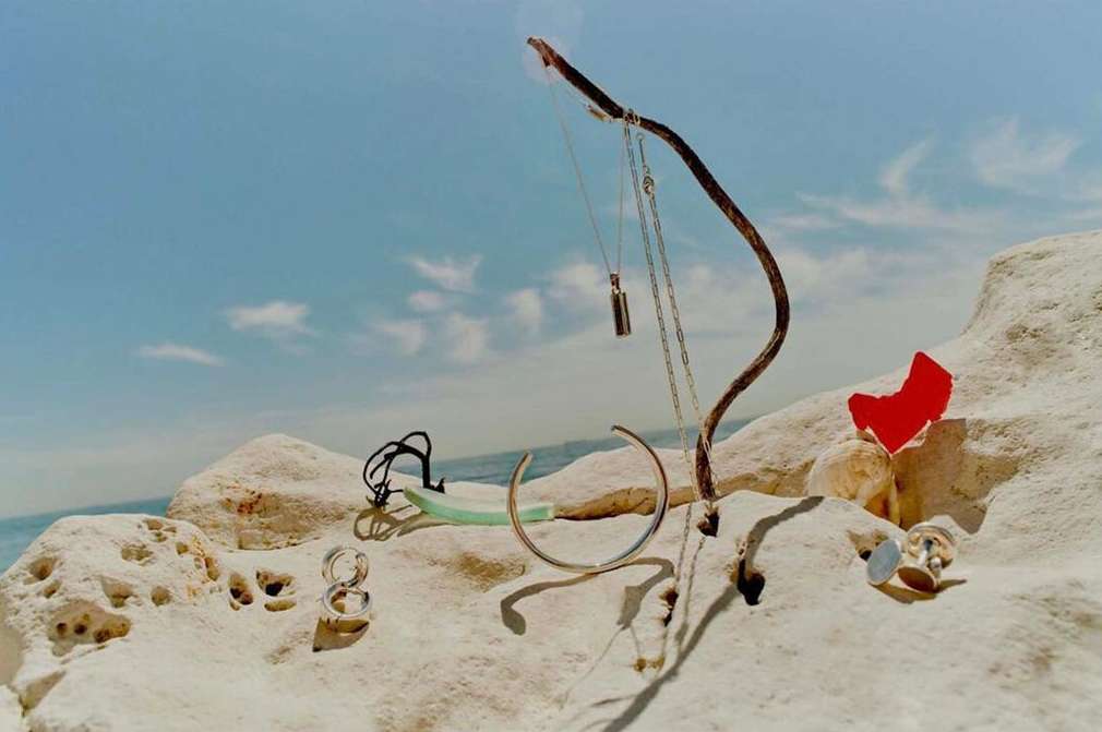 Still life jewelry display featuring necklaces, bracelets, and rings arranged on rocks at the beach with a cloudy blue sky background.