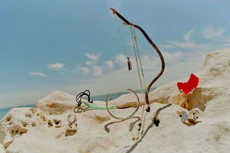 Still life jewelry display featuring necklaces, bracelets, and rings arranged on rocks at the beach with a cloudy blue sky background.