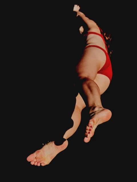 A person in a red swimsuit with arms extended in a dark indoor space, showing their full body and bare feet.