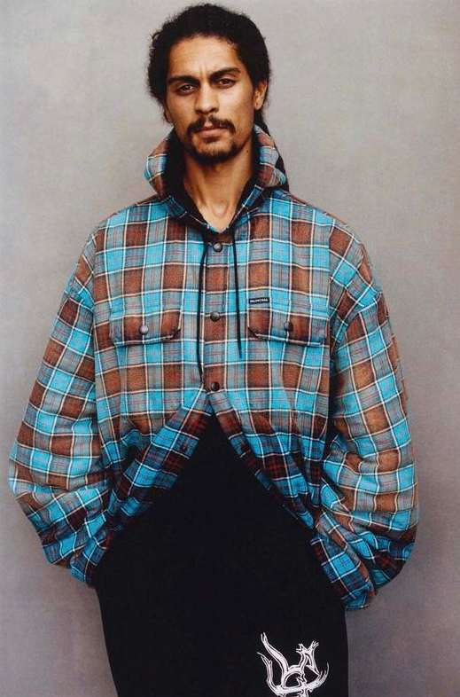 Adult male posing in a hooded plaid shirt and black sweatpants against a neutral gray studio backdrop, looking directly at the camera.