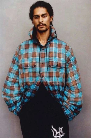 Adult male posing in a hooded plaid shirt and black sweatpants against a neutral gray studio backdrop, looking directly at the camera.