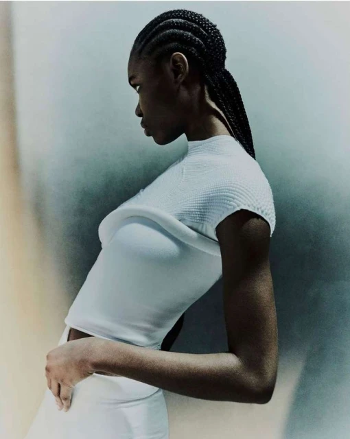 A young Black woman with braided hair wears an all-white outfit against a textured wall, looking to the left in a fashion-editorial style.