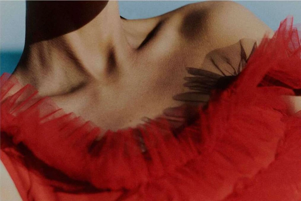Upper body shot of an adult female wearing red tulle fabric, illuminated by natural light, against a soft blue background, studio lighting.