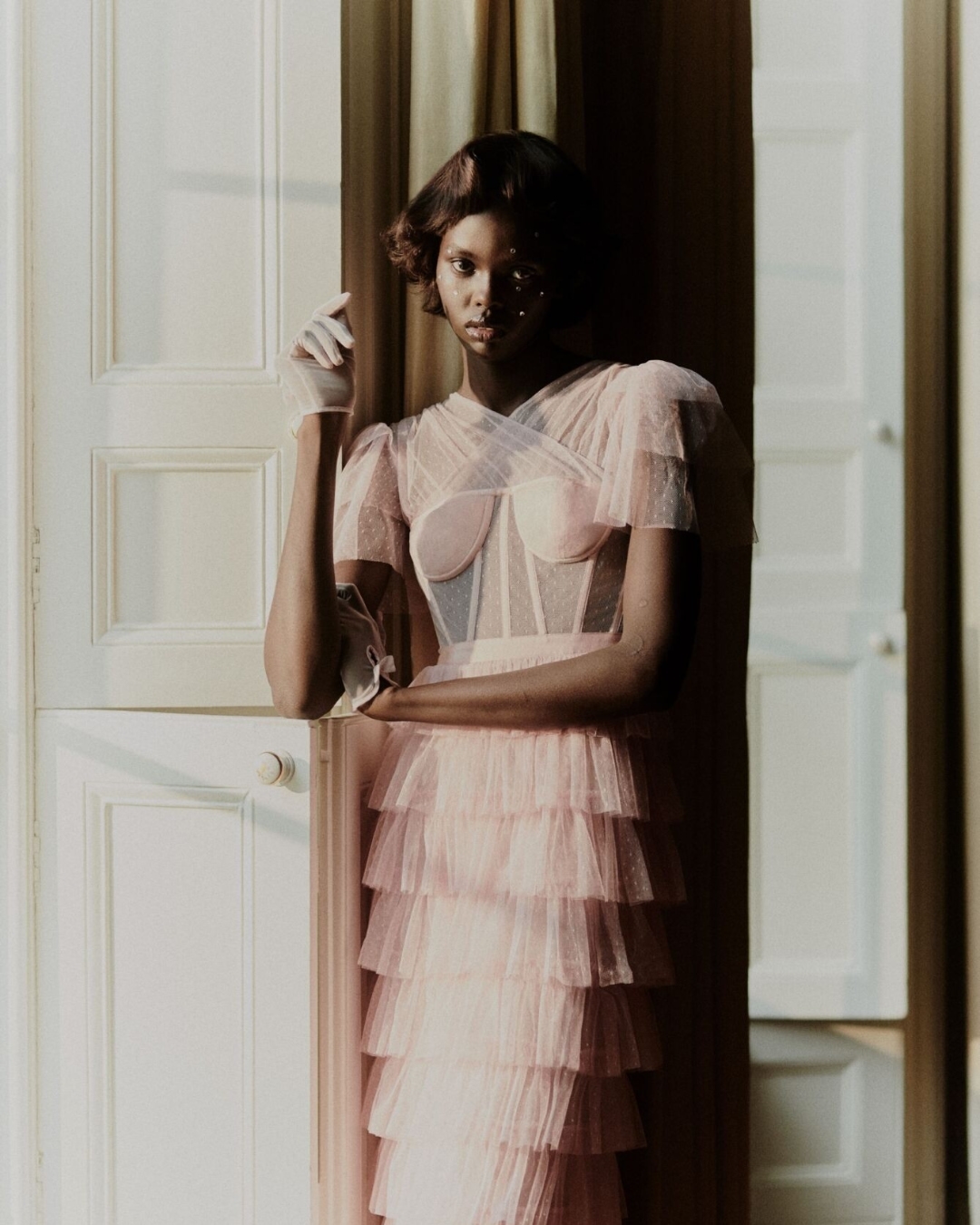 Elegant African American woman in a pink tulle dress poses in front of a white door with natural light illuminating the scene indoors.