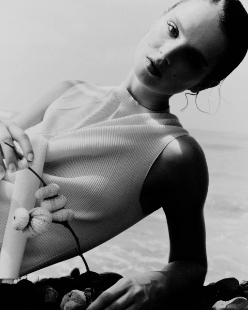 Monochrome image of a woman lying on a beach holding stacked shells. She has dark hair and is wearing a ribbed top.