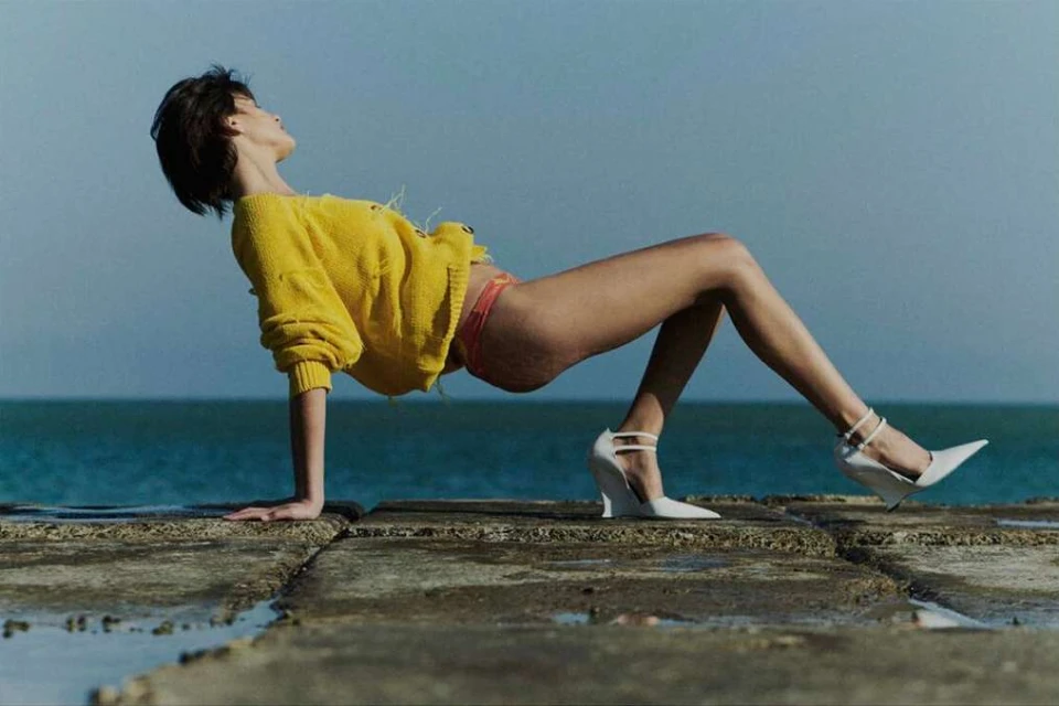 Fashion shot featuring a woman in a yellow sweater and heels posing on stone blocks near the ocean under a clear sky.
