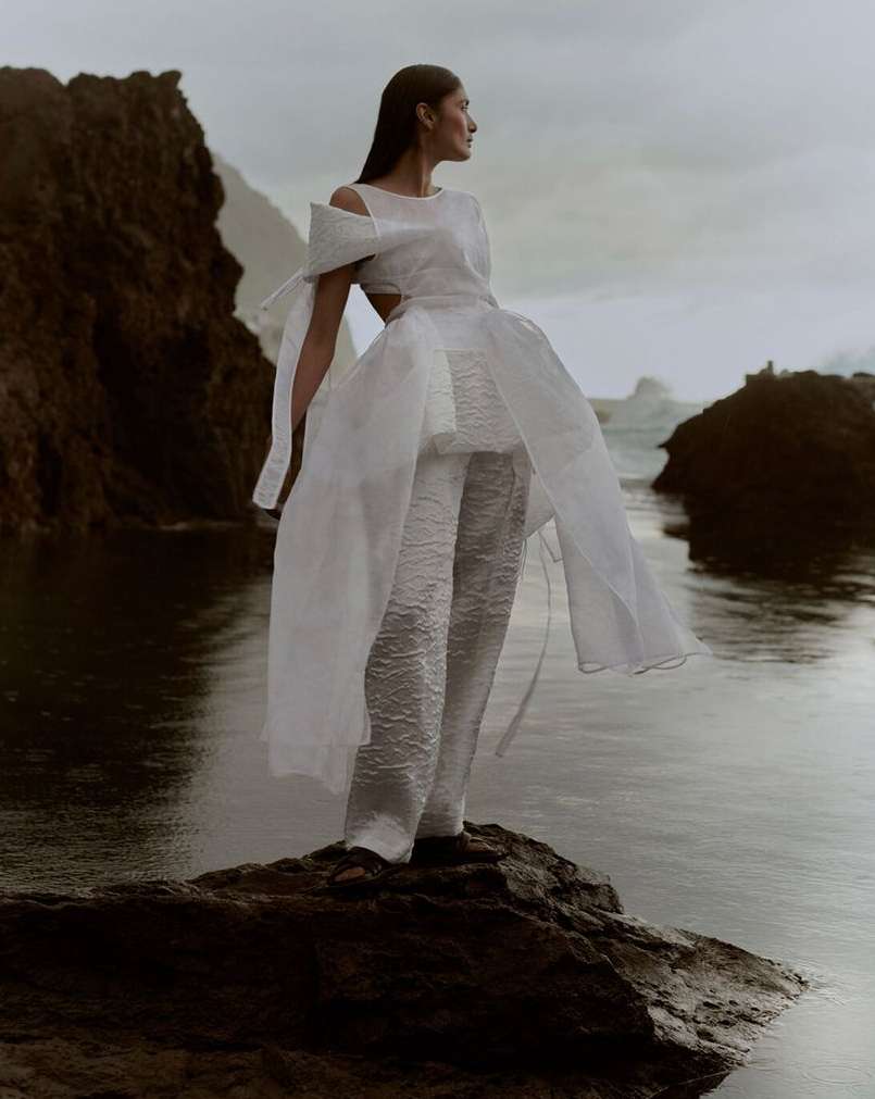 Woman in a white dress and pants stands on a rock formation by the sea, gazing into the distance with ocean and rock formations in the background.