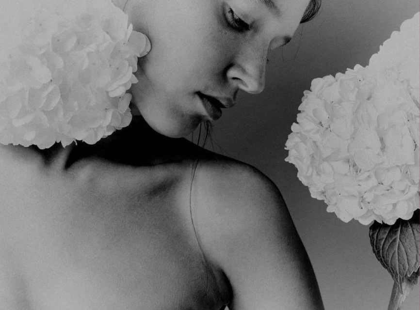 Black-and-white close-up of a young woman's face and shoulders, framed by large, white hydrangea blooms. Minimalist beauty portrait.