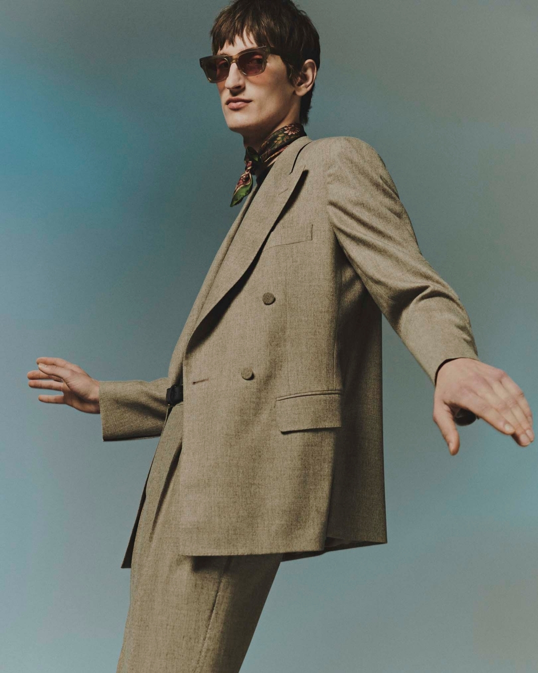 Man wearing sunglasses and a patterned scarf with a tan double-breasted suit against a gradient background.