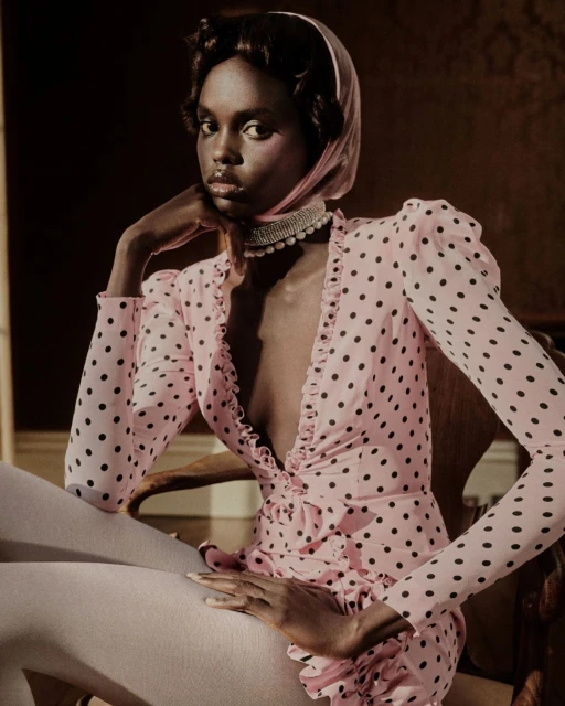 Elegant Black woman poses in a pink polka dot dress and headscarf, seated in a vintage chair, with a dark neutral backdrop in a fashion portrait.