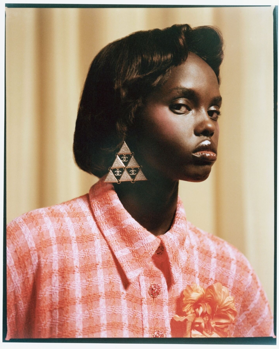 Elegant, dark-skinned woman in a coral-colored jacket and triangular earrings featuring a Chanel logo, posing against a tan background.
