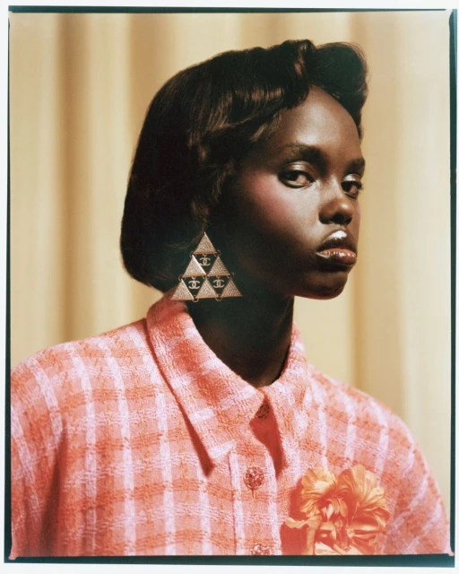 Elegant, dark-skinned woman in a coral-colored jacket and triangular earrings featuring a Chanel logo, posing against a tan background.