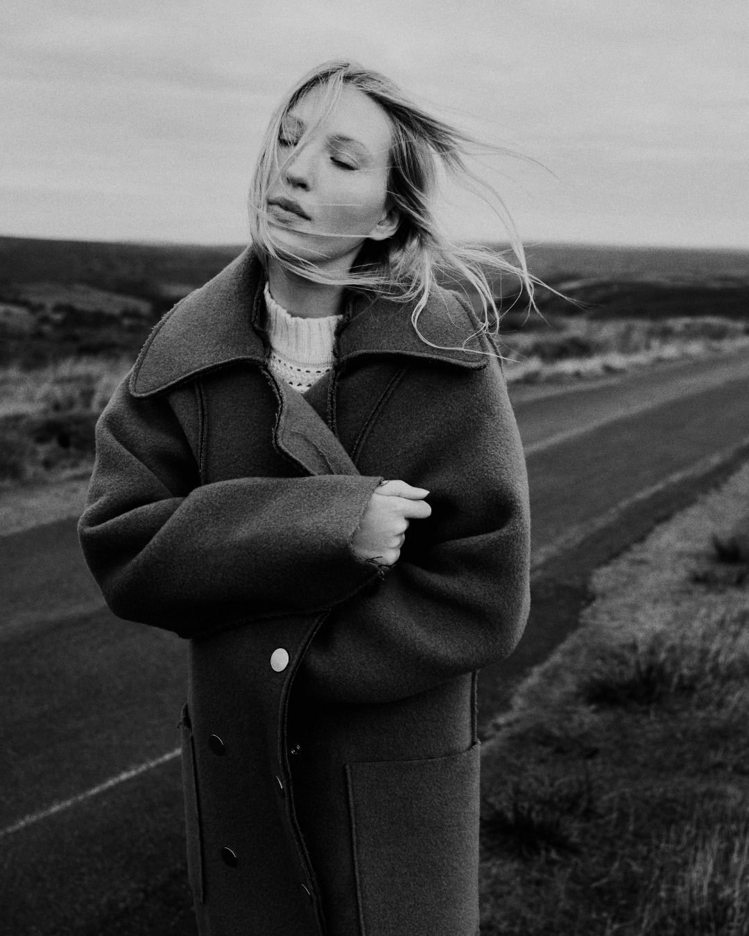 Monochrome portrait of a woman with long blonde hair wearing a heavy coat standing outdoors on a rural road