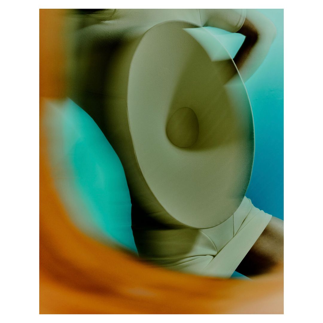 Abstract fashion shot featuring a person in a tan outfit behind a circular object with blurred orange and blue background.