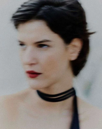 Close-up portrait of a woman with short dark hair wearing a black choker and red lipstick against a neutral background.