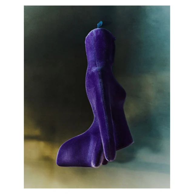 Abstract purple velvet sculpture against a mottled green and dark grey background with a single, small blue feather accent.