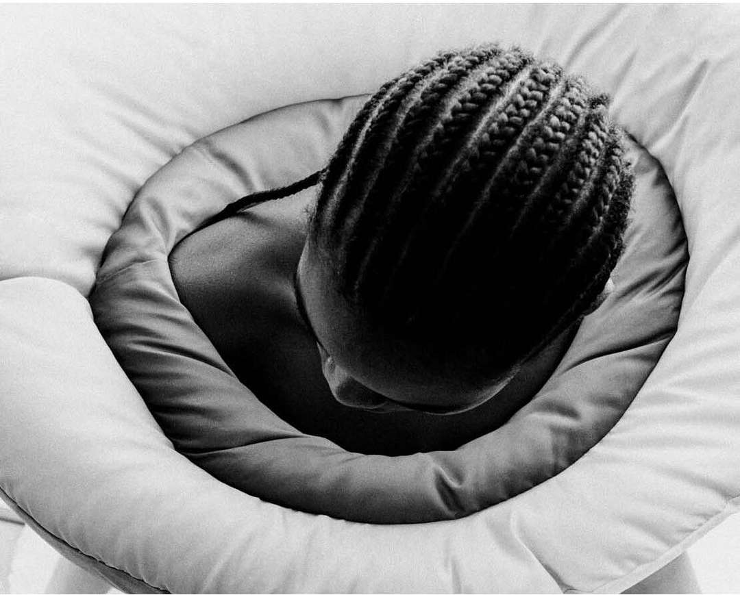 Black and white close-up of a person with cornrow braids framed by a padded, circular structure, creating a soft, intimate portrait.