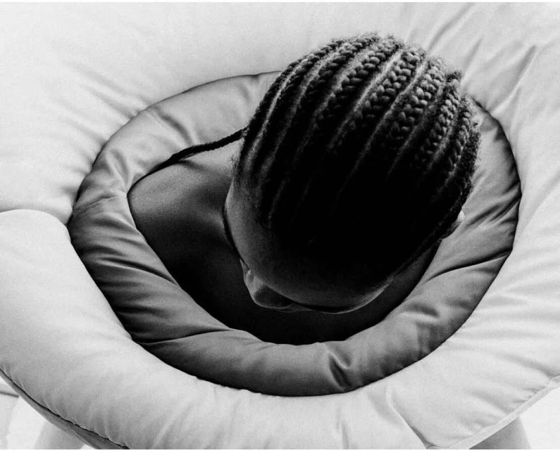 Black and white close-up of a person with cornrow braids framed by a padded, circular structure, creating a soft, intimate portrait.