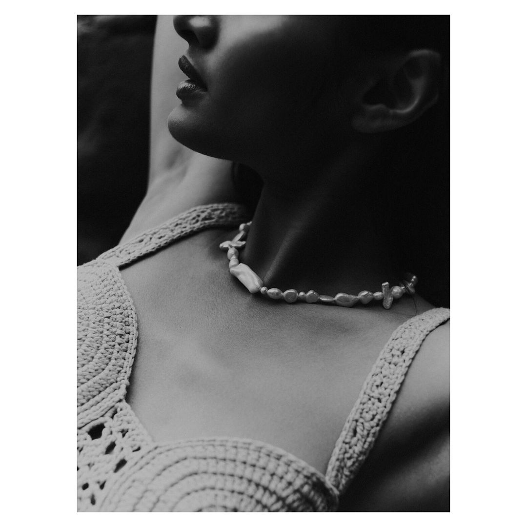 Monochrome portrait of a woman with a pearl necklace, wearing a crochet top, photographed in soft lighting, evoking natural beauty.