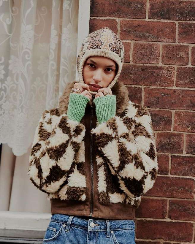 Woman in patterned hat and jacket posing by a brick wall with a window, styled in a fashion-forward and urban setting.