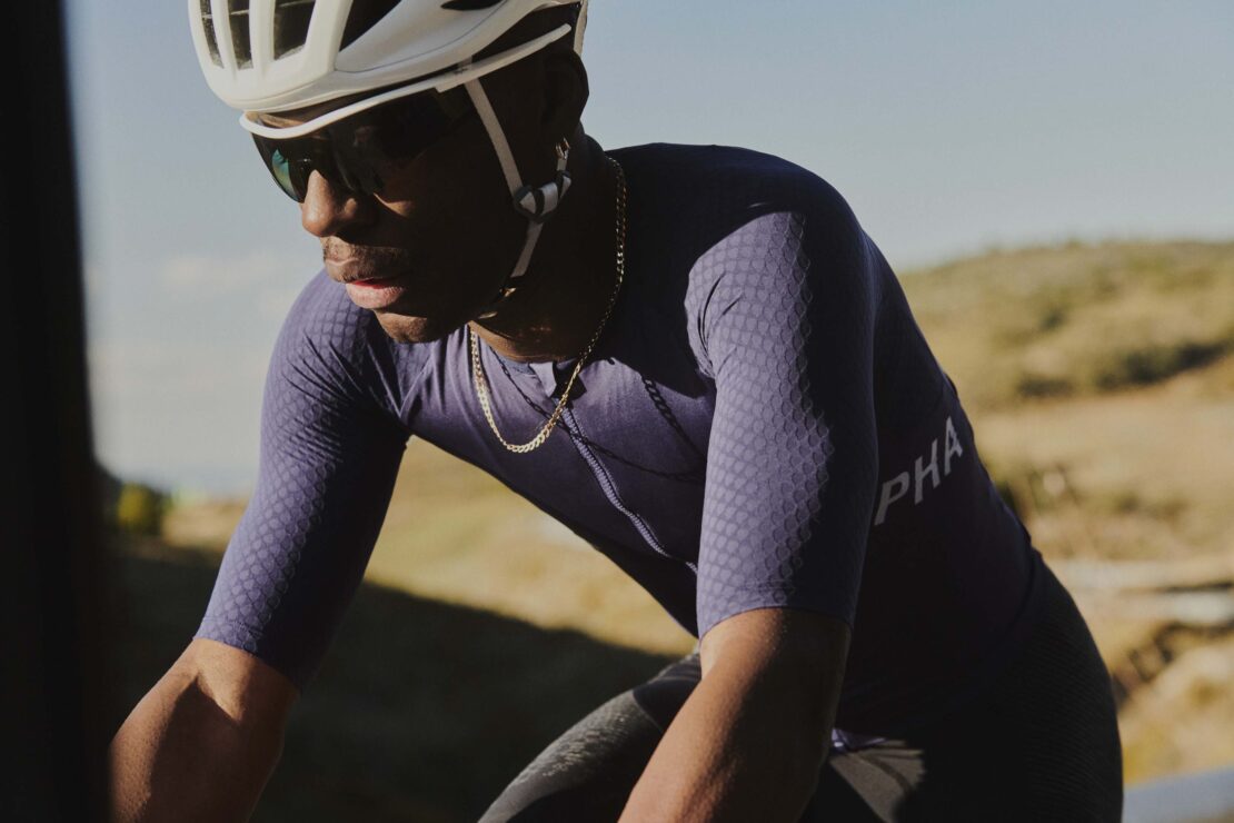 An African-American male cyclist rides his bicycle along a road with hills in the background, wearing a helmet and sunglasses.