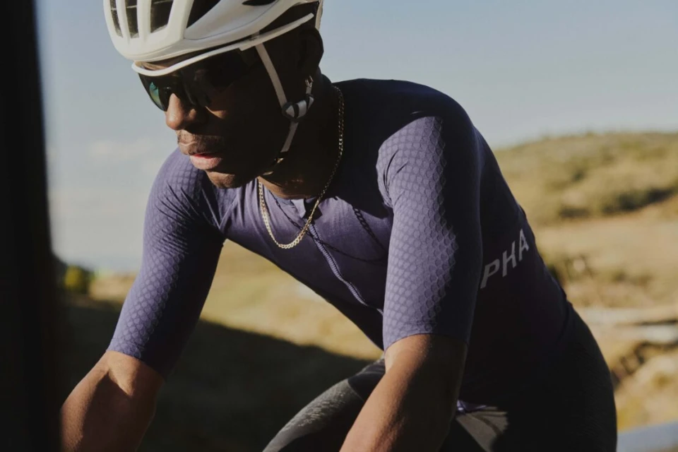 An African-American male cyclist rides his bicycle along a road with hills in the background, wearing a helmet and sunglasses.