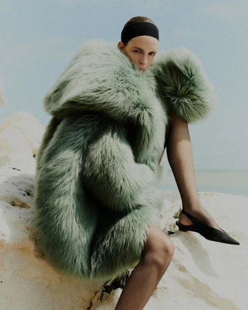 Model in a voluminous green faux fur hooded coat and black slingback shoes seated against white rocks under a clear sky