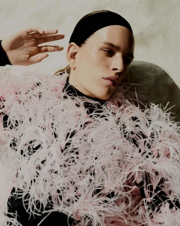 Model wearing a textured pink feather jacket and black headband against a neutral textured stone background by Daniel Benson