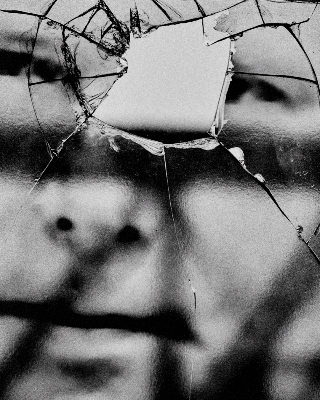 Monochrome portrait showing a partial human face viewed through a shattered glass surface, creating a distorted, fragmented image.