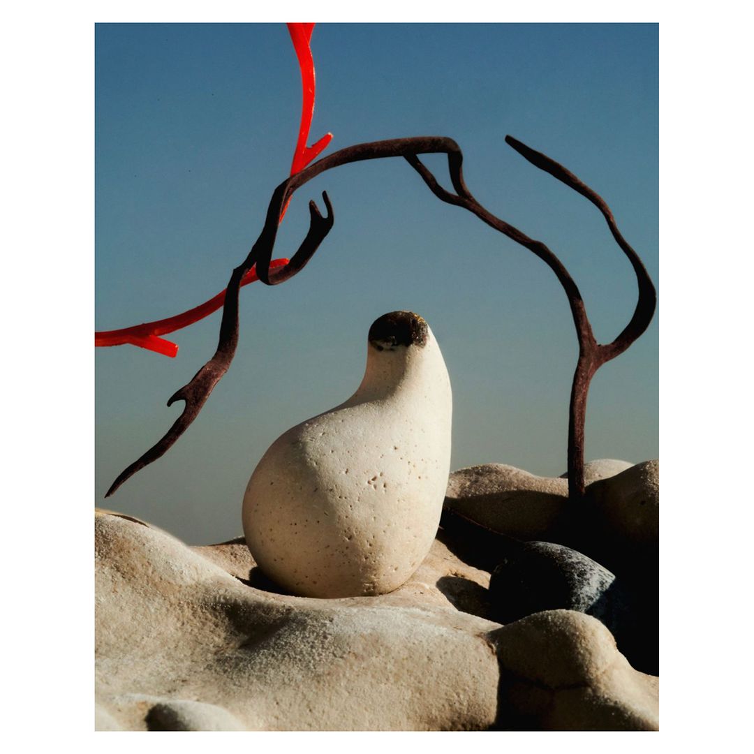 Abstract still life featuring a smooth, beige stone sculpture on a rock formation, accented by curved, painted branches against a light blue sky.