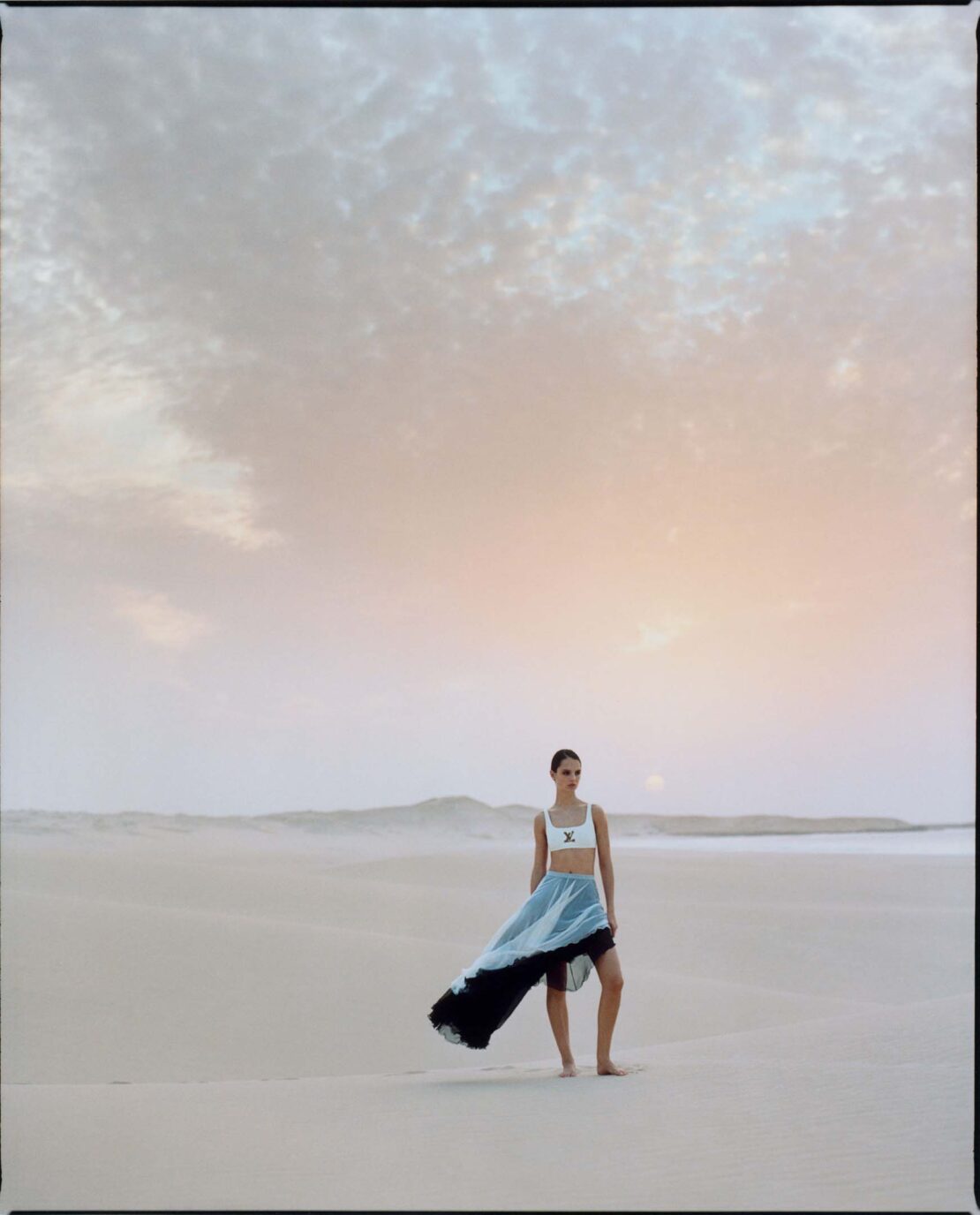 Fashion shot of a model in a light-colored top and long, flowing skirt posing on a sandy beach beneath a cloudy sky at dusk.