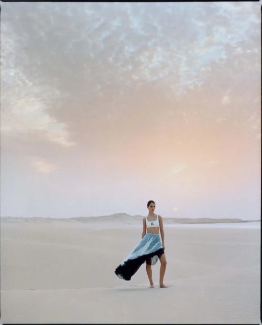 Fashion shot of a model in a light-colored top and long, flowing skirt posing on a sandy beach beneath a cloudy sky at dusk.