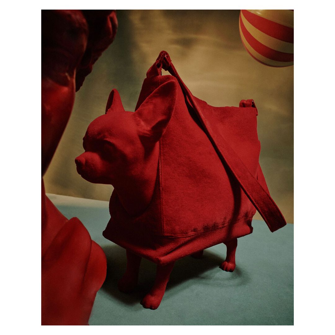 Red dog sculpture appears to carry a matching red tote bag, set against a sky-like backdrop and a spherical striped prop.