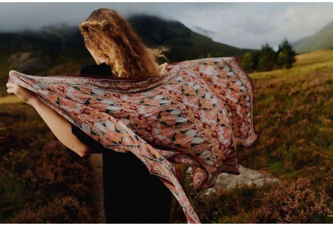 Back view of adult female with wavy hair holding a patterned scarf blowing in the wind in a grassy highland landscape.