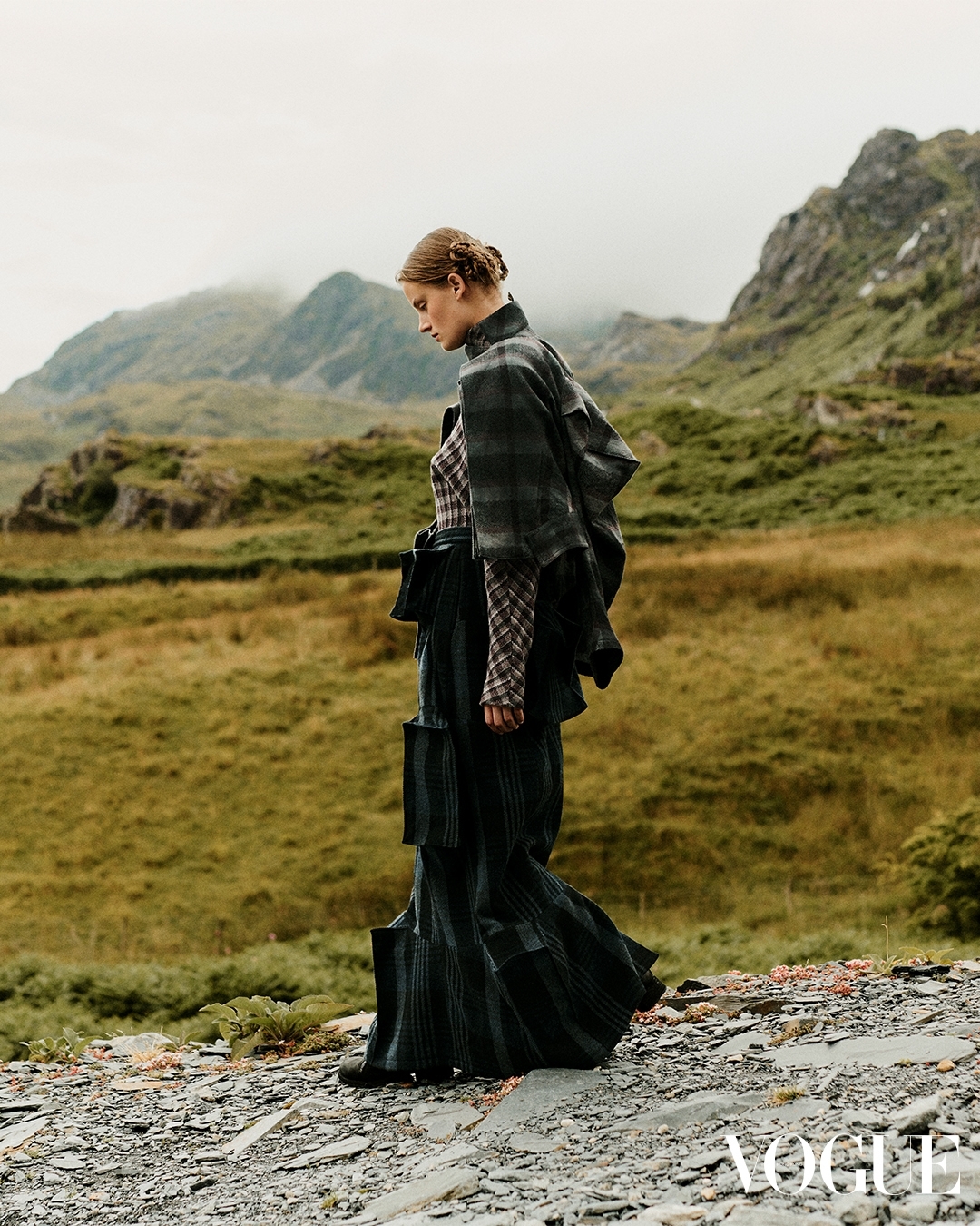 A fashion shot features a woman in a plaid outfit standing in a mountain landscape under an overcast sky, showcasing editorial style.