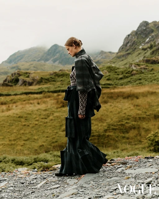 A fashion shot features a woman in a plaid outfit standing in a mountain landscape under an overcast sky, showcasing editorial style.