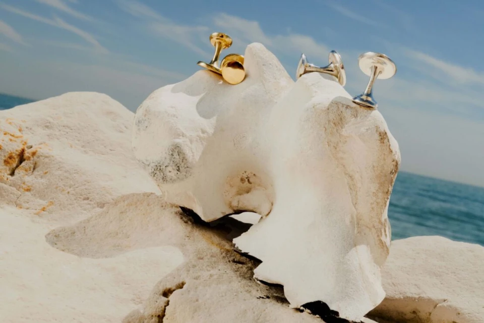 Two metal cufflinks in gold and silver displayed on white textured stones with a background of blue ocean water and sky.
