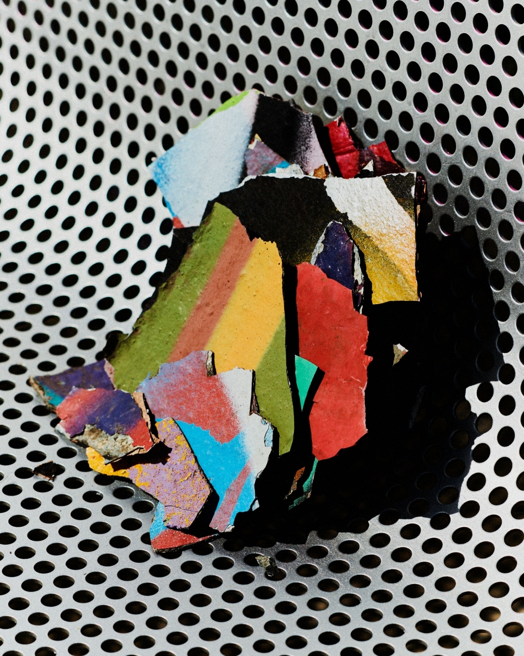 Close-up photograph of fragmented, colorful paint chips arranged on a textured, perforated metal surface with contrasting light and shadows.