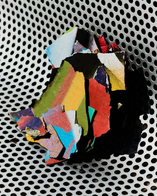 Close-up photograph of fragmented, colorful paint chips arranged on a textured, perforated metal surface with contrasting light and shadows.
