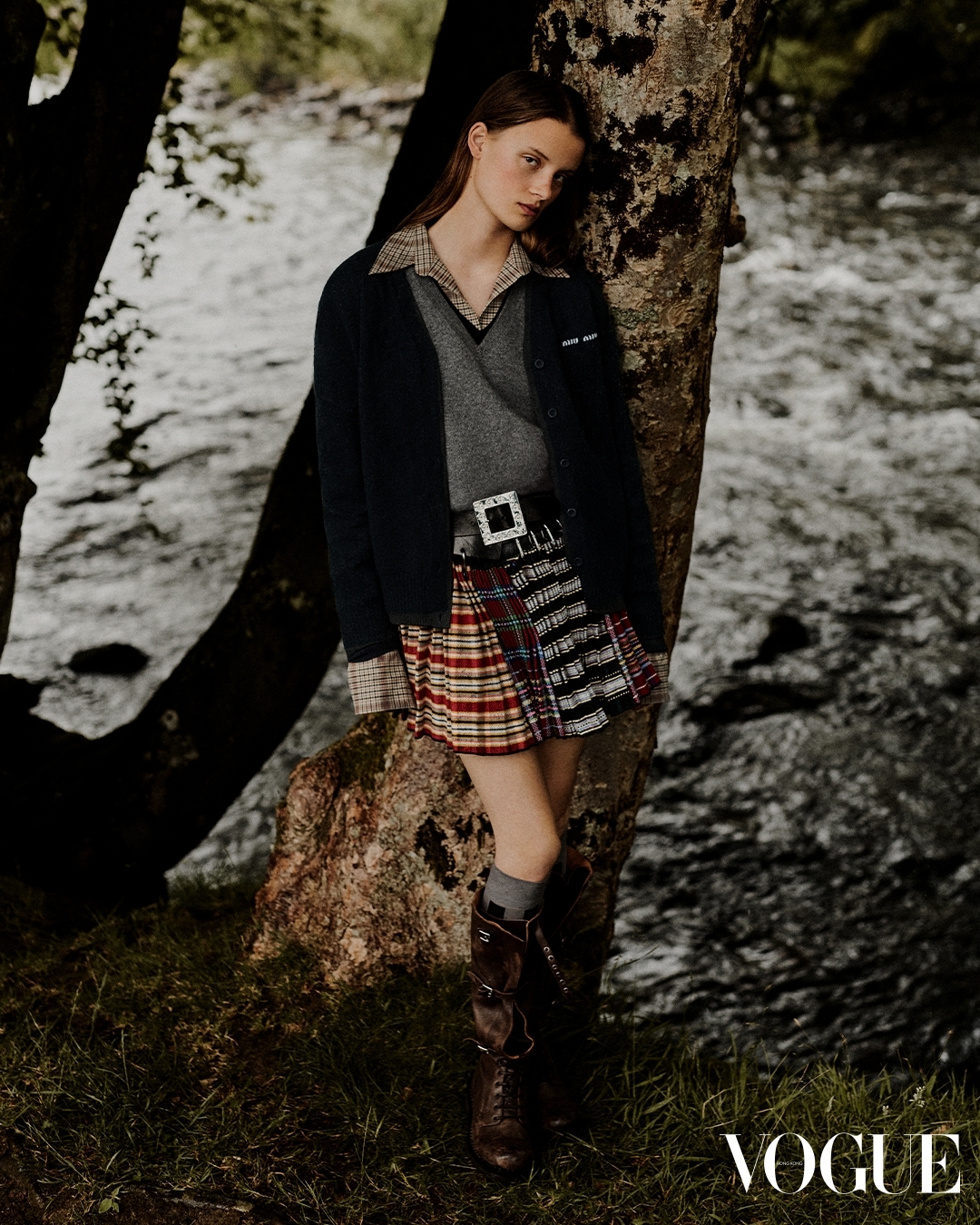 Young woman in tartan skirt and brown boots leaning against a tree by a flowing river. Moody outdoor fashion shot. Vogue logo visible.