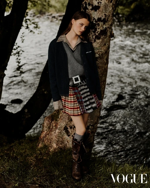 Young woman in tartan skirt and brown boots leaning against a tree by a flowing river. Moody outdoor fashion shot. Vogue logo visible.
