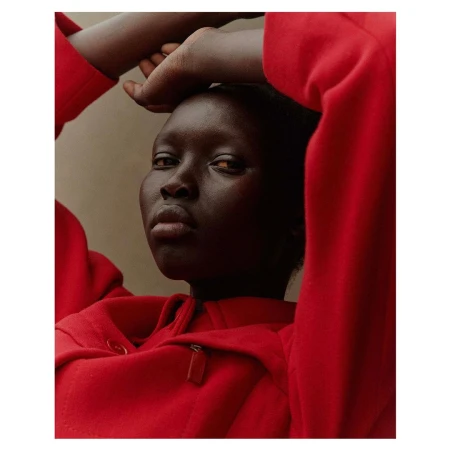 Portrait of a Black woman in a vibrant red hoodie with hands raised behind her head against a neutral wall