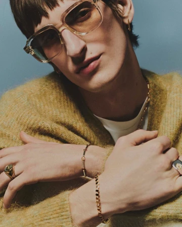 Close-up portrait of a man wearing tinted oversized glasses, a fuzzy mustard sweater, and gold chains and rings photographed by Daniel Benson.