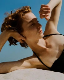 Close-up portrait of a red-haired woman with freckles lying against a light sandy surface under a clear blue sky