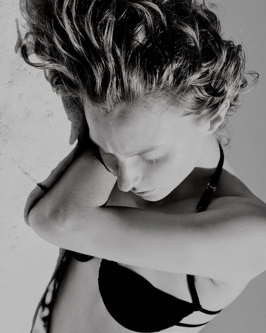 High-angle black and white portrait of a woman with curly hair resting her head on her crossed arms, wearing a black strap top.