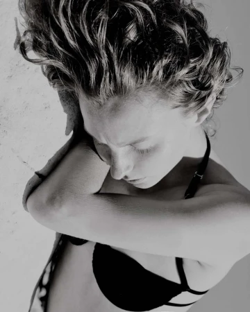 High-angle black and white portrait of a woman with curly hair resting her head on her crossed arms, wearing a black strap top.