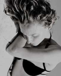 High-angle black and white portrait of a woman with curly hair resting her head on her crossed arms, wearing a black strap top.